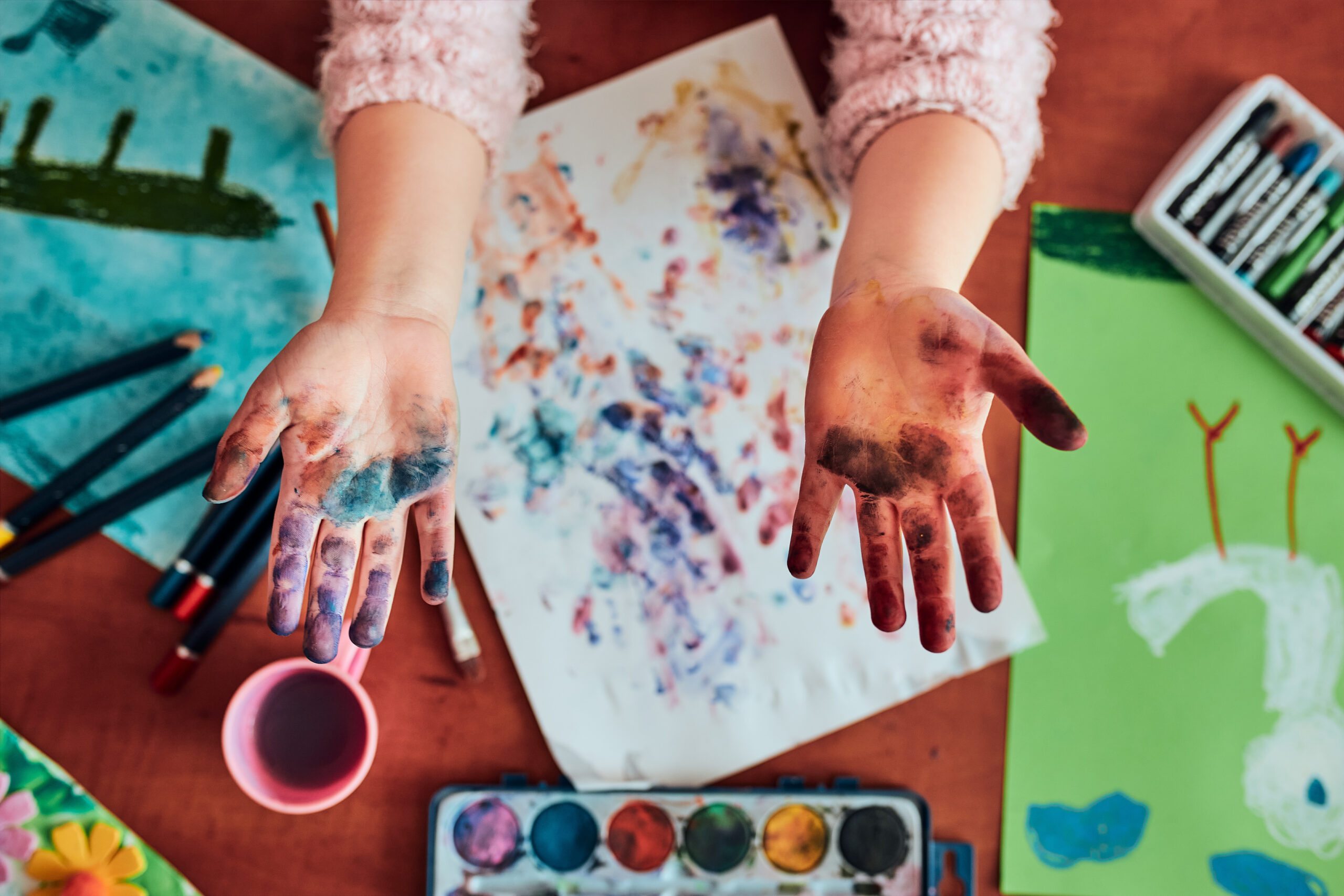 Little girl preschooler showing painted colourful hands. Child having fun making a stamps on sheet of paper with painted hands during an art class in the classroom